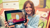 Mädchen hält ein Tablet mit laufendem KatzeQ-Spiel und lächelt in die Kamera, Release-Pressefoto.